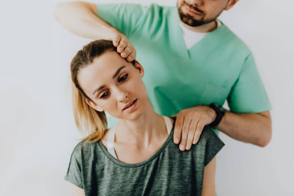 A woman sits calmly and side bending her neck as a physiotherapist massages her shoulder and head in a bright clinic.