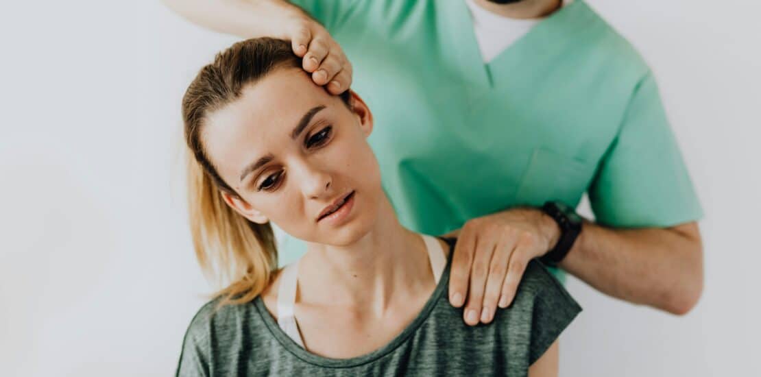 A woman sits calmly and side bending her neck as a physiotherapist massages her shoulder and head in a bright clinic.