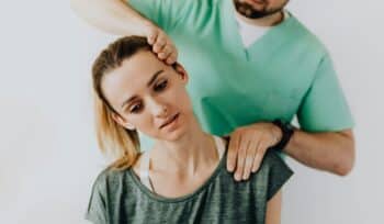 A woman sits calmly and side bending her neck as a physiotherapist massages her shoulder and head in a bright clinic.