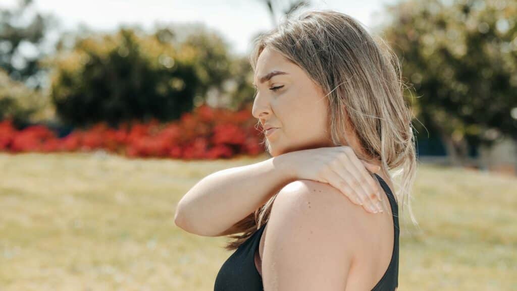 A woman in workout attire pauses her park exercise, touching her shoulder in pain.