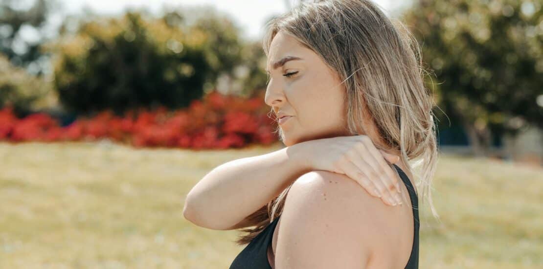 A woman in workout attire pauses her park exercise, touching her shoulder in pain.