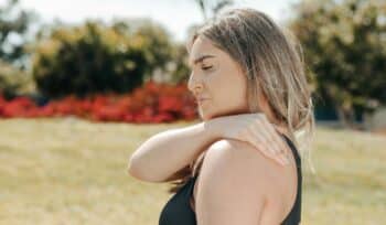 A woman in workout attire pauses her park exercise, touching her shoulder in pain.