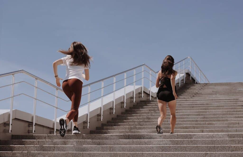 Two women are running up a set of stairs together, both appearing energetic and focused.