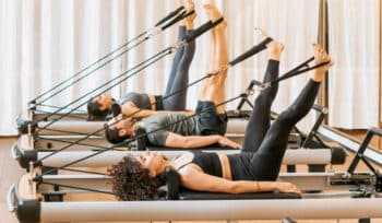 Women performing Pilates exercises on reformer machines, using resistance bands to stretch their legs upward.