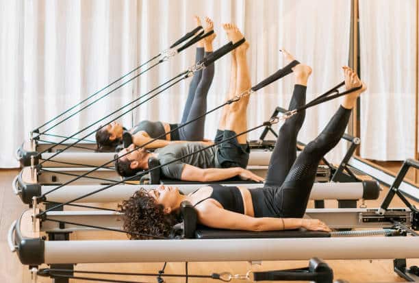 Women performing Pilates exercises on reformer machines, using resistance bands to stretch their legs upward.