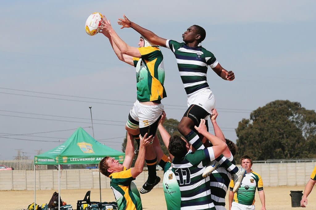 Two AFL players contesting the ball in mid-air, surrounded by four opponents, grabbing their hamstring. The intense action captures the competitive spirit of Australian football league gameplay.