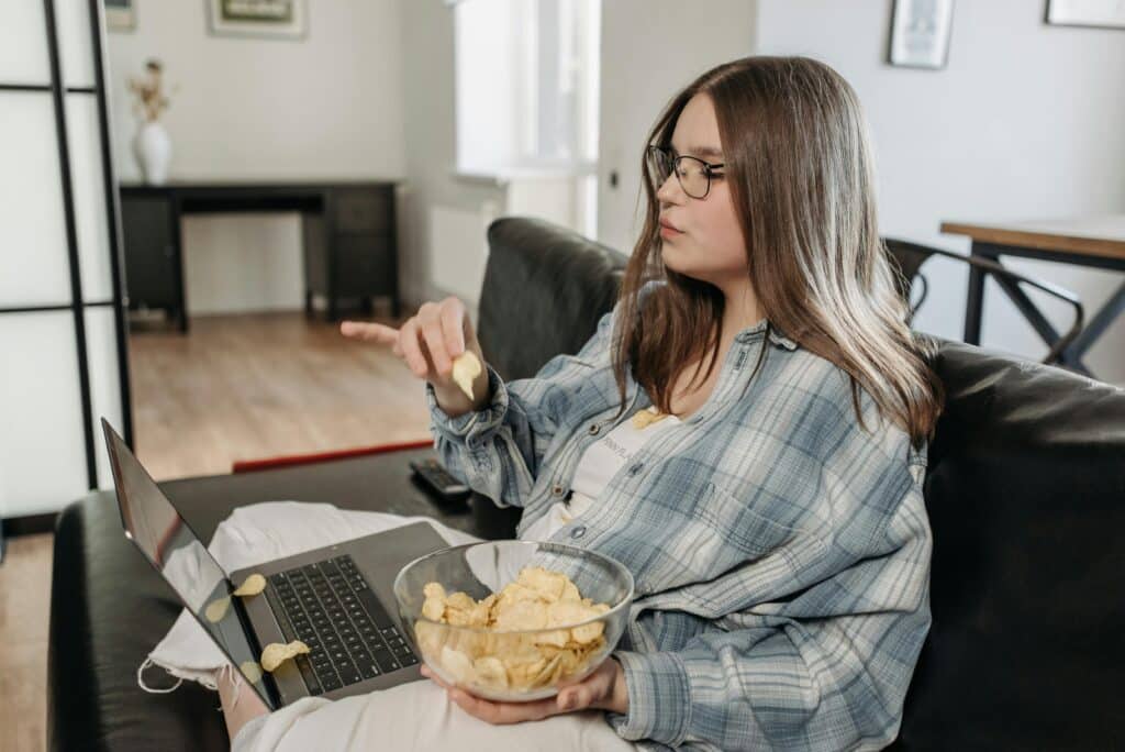 A woman leads a sedentary lifestyle as she sits on a sofa, using her laptop and eating potato chips, with some chips spilled on the laptop.