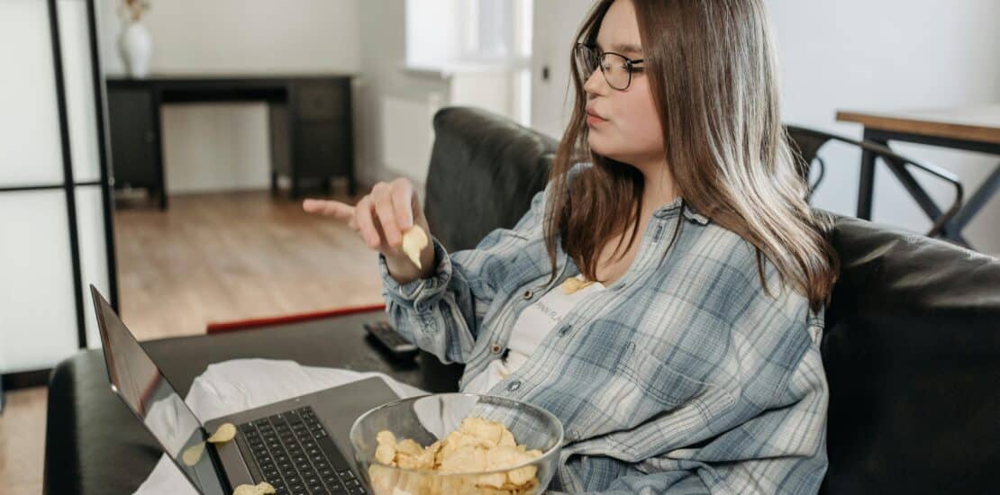 A woman leads a sedentary lifestyle as she sits on a sofa, using her laptop and eating potato chips, with some chips spilled on the laptop.