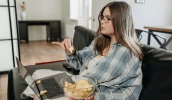 A woman leads a sedentary lifestyle as she sits on a sofa, using her laptop and eating potato chips, with some chips spilled on the laptop.