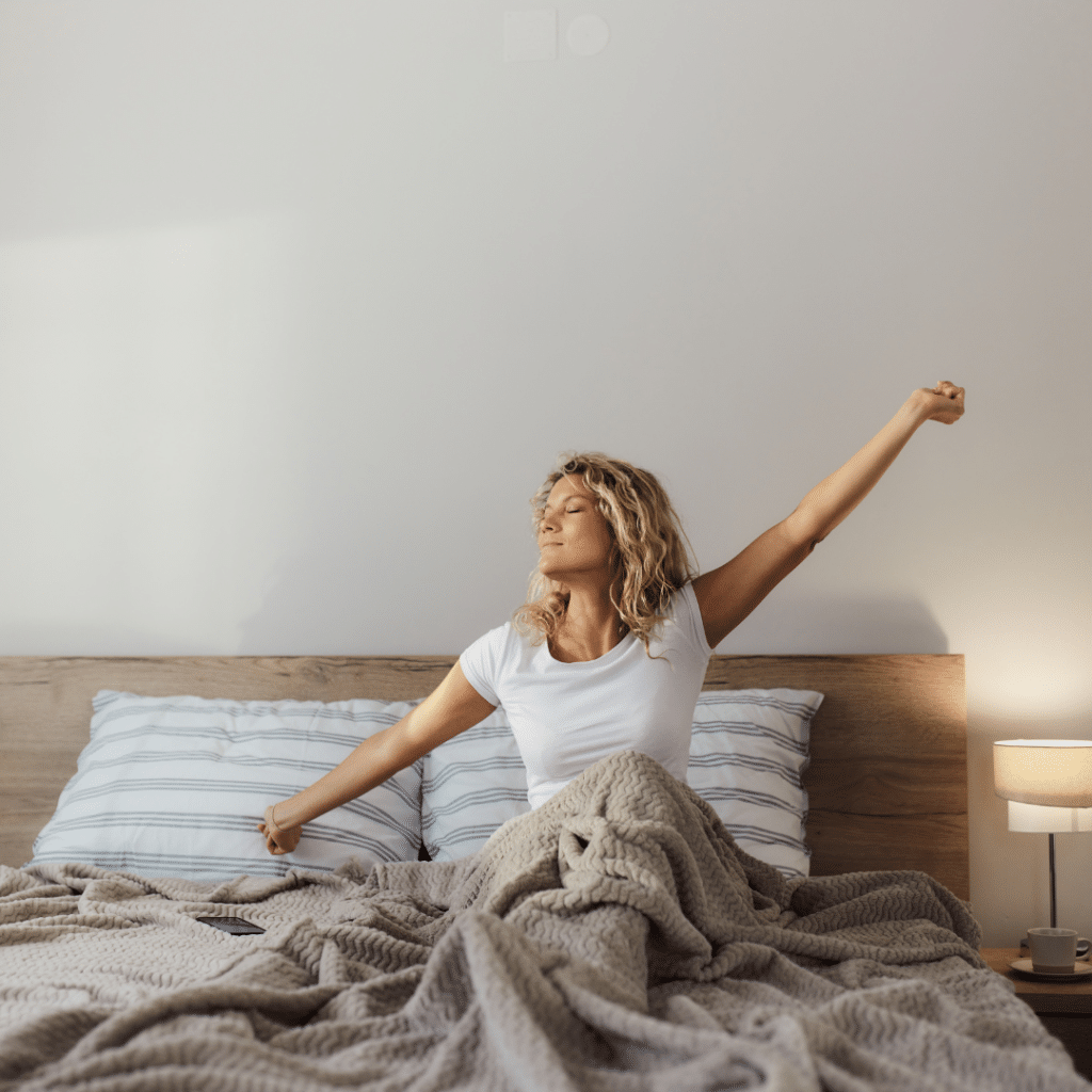 A woman in cozy pajamas sits up in a comfortable-looking bed, just waking up, looking happy, fulfilled and refreshed