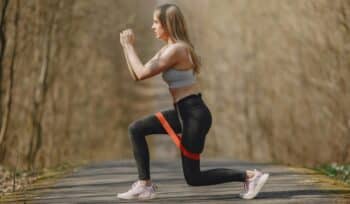 A woman is exercising in the park, using a resistance band around her knees. performing a lateral band walk to strengthen her leg muscles. She looks focused and determined, wearing athletic clothing and sneakers.