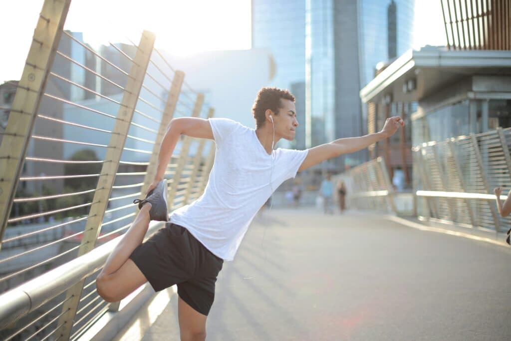 A man in a white shirt performs leg stretches while leaning on a railing by the bay. Enjoying outdoor exercise and stretching for fitness and well-being.