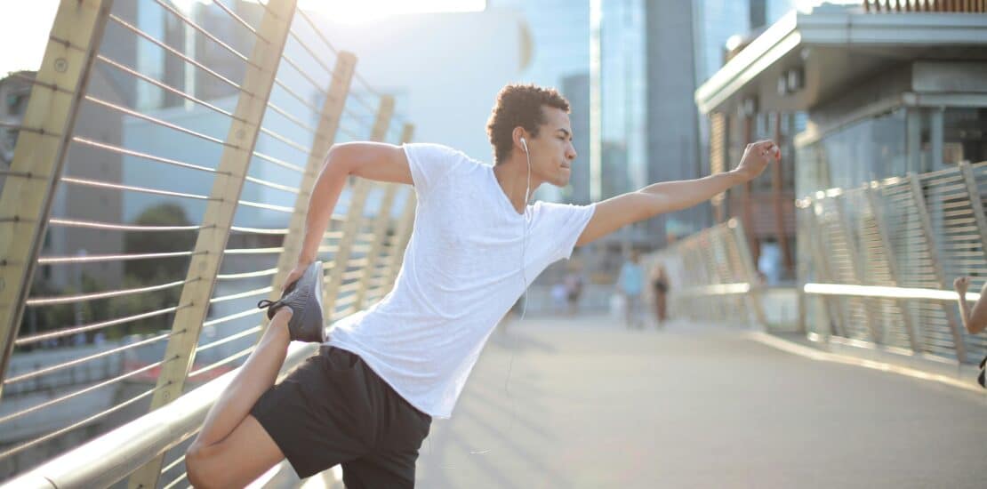 A man in a white shirt performs leg stretches while leaning on a railing by the bay. Enjoying outdoor exercise and stretching for fitness and well-being.