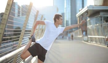 A man in a white shirt performs leg stretches while leaning on a railing by the bay. Enjoying outdoor exercise and stretching for fitness and well-being.