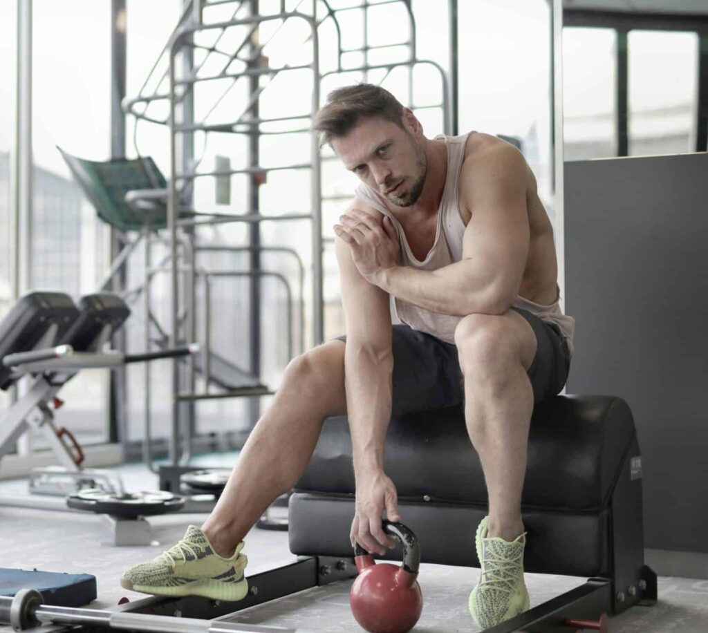 A man working out in the gym, touching his shoulder possibly due to shoulder bursitis, while exercising with a kettlebell.