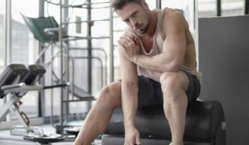 A man working out in the gym, touching his shoulder possibly due to shoulder bursitis, while exercising with a kettlebell.