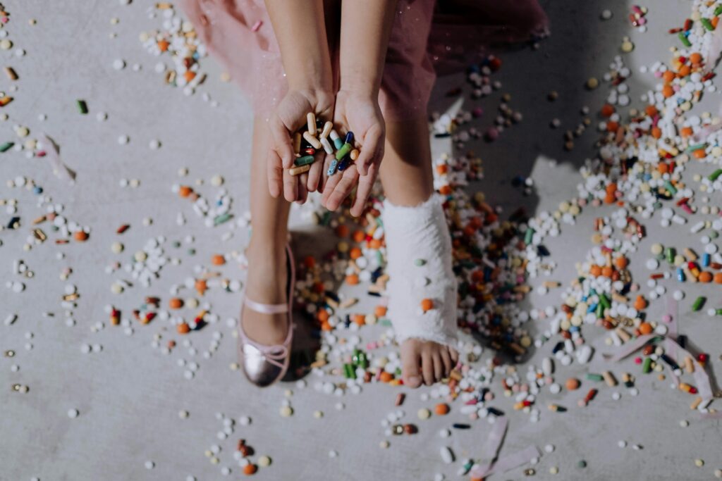 Image of a ballerina's feet: one foot is wrapped in a medical bandage for an injury, while the other foot is wearing a ballet shoe. The ballerina is holding a handful of medication pills, with additional pills scattered across the floor.