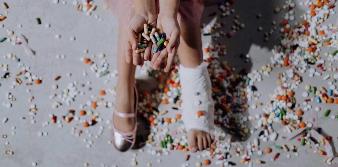 Image of a ballerina's feet: one foot is wrapped in a medical bandage for an injury, while the other foot is wearing a ballet shoe. The ballerina is holding a handful of medication pills, with additional pills scattered across the floor.