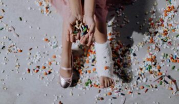 Image of a ballerina's feet: one foot is wrapped in a medical bandage for an injury, while the other foot is wearing a ballet shoe. The ballerina is holding a handful of medication pills, with additional pills scattered across the floor.