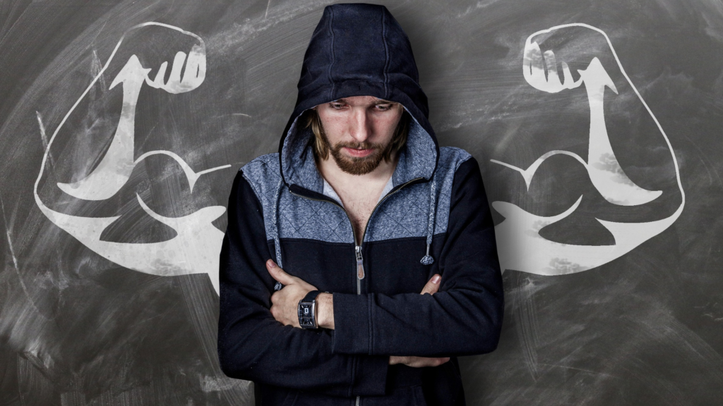 A man in a jacket, hiding or wrapping his arm around his body and looking down, appears embarrassed. Behind him is a chalk drawing of muscular arms, highlighting the contrast and foreshadowing the effects of sarcopenia.