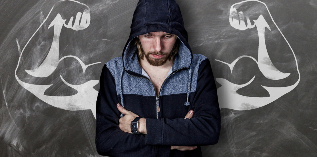 A man in a jacket, hiding or wrapping his arm around his body and looking down, appears embarrassed. Behind him is a chalk drawing of muscular arms, highlighting the contrast and foreshadowing the effects of sarcopenia.