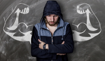 A man in a jacket, hiding or wrapping his arm around his body and looking down, appears embarrassed. Behind him is a chalk drawing of muscular arms, highlighting the contrast and foreshadowing the effects of sarcopenia.