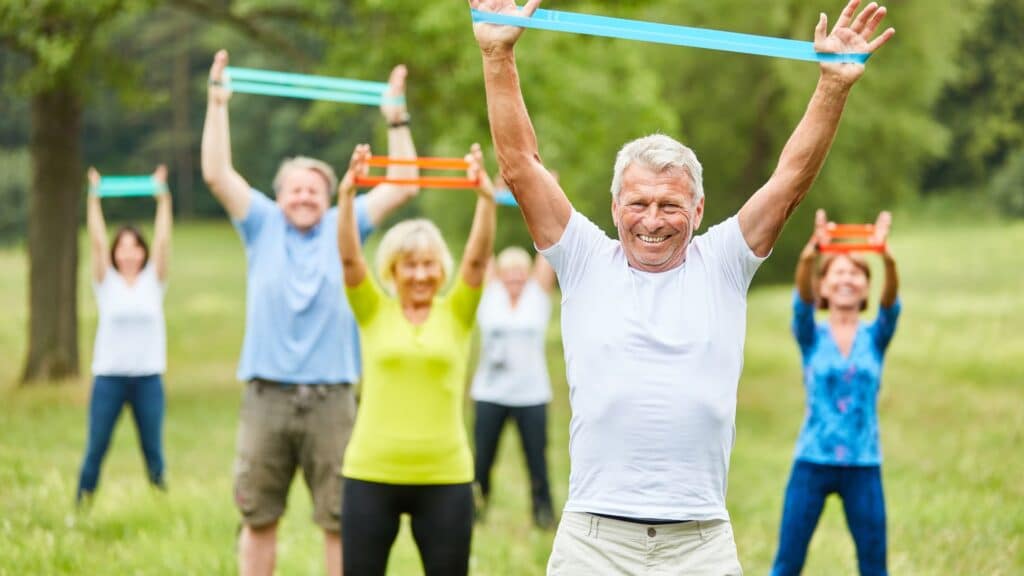 A group of elderly individuals exercising outdoors in a park, using resistance bands in both hands. They appear engaged and focused, with green trees and a bright sky in the background, promoting a vibrant and active lifestyle.