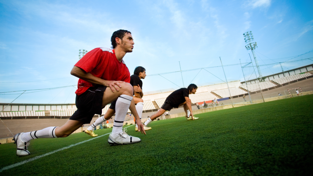 A group of athletes doing warm exercises in an open stadium, stretching their legs with one leg bent, preparing for training.