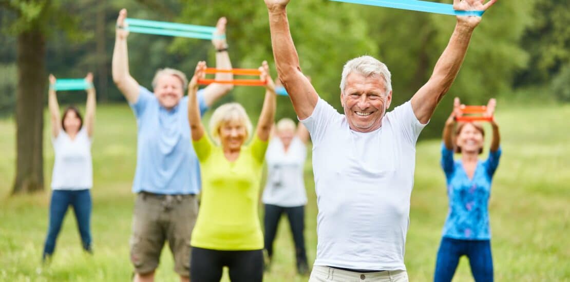 A group of elderly individuals exercising outdoors in a park, using resistance bands in both hands. They appear engaged and focused, with green trees and a bright sky in the background, promoting a vibrant and active lifestyle.