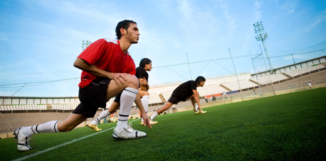 A group of athletes doing warm exercises in an open stadium, stretching their legs with one leg bent, preparing for training.