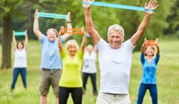 A group of elderly individuals exercising outdoors in a park, using resistance bands in both hands. They appear engaged and focused, with green trees and a bright sky in the background, promoting a vibrant and active lifestyle.