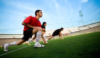 A group of athletes doing warm exercises in an open stadium, stretching their legs with one leg bent, preparing for training.