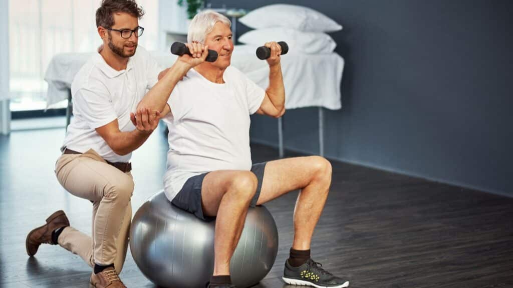 An older man sits on an exercise ball, lifting small weights in each hand while being assisted by a physiotherapist, focusing on core strength.