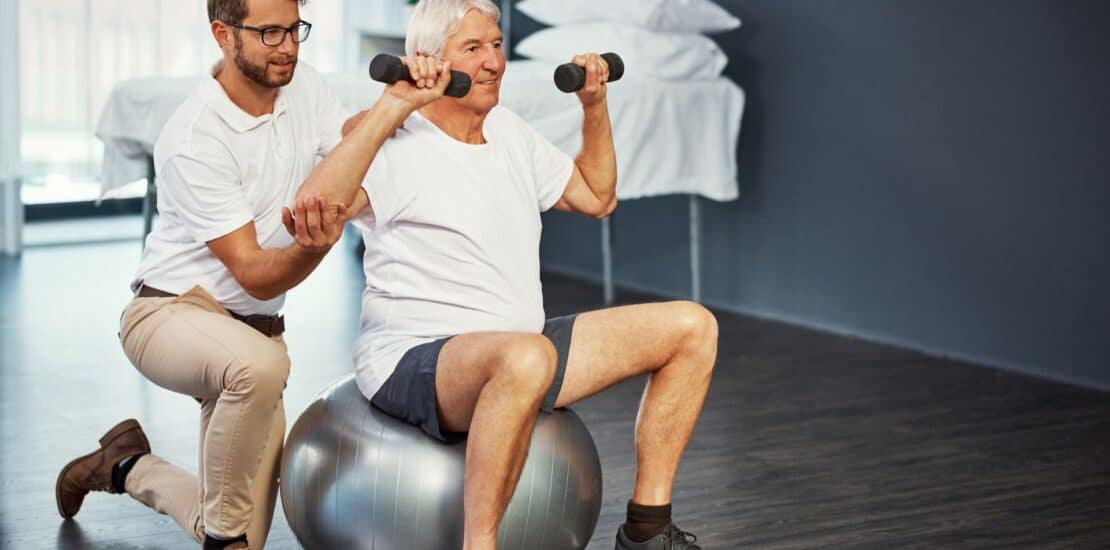 An older man sits on an exercise ball, lifting small weights in each hand while being assisted by a physiotherapist, focusing on core strength.