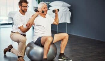 An older man sits on an exercise ball, lifting small weights in each hand while being assisted by a physiotherapist, focusing on core strength.