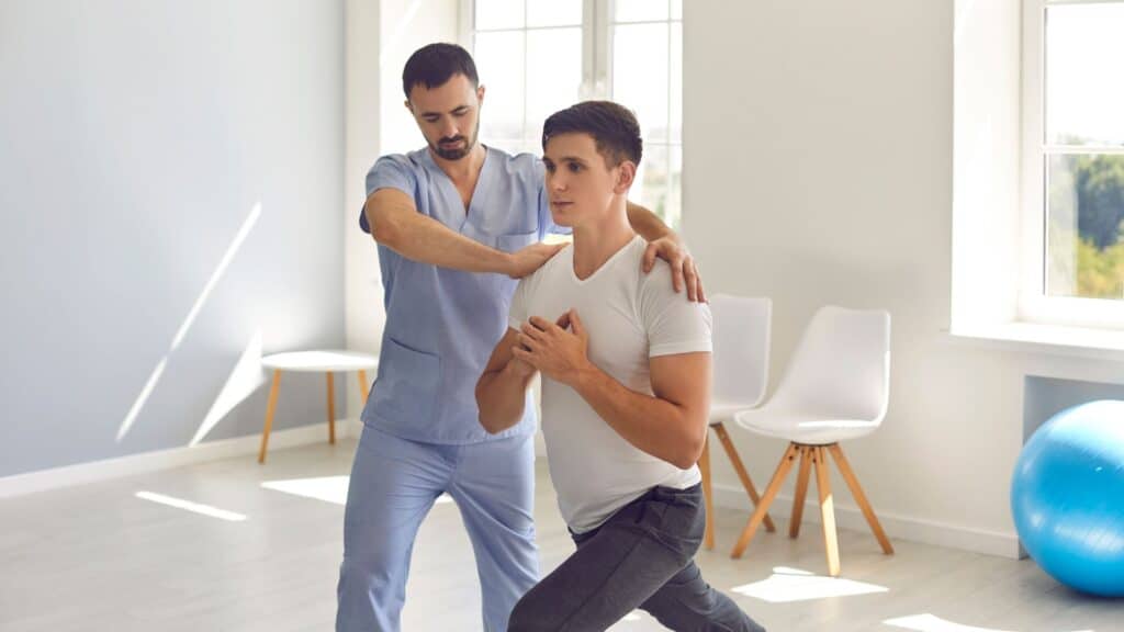 A patient is half-bending one leg while bringing his hands together at his core, demonstrating focus and balance. A physiotherapist stands beside him, providing guidance and support in a well-equipped physiotherapy clinic. The atmosphere is encouraging and professional, highlighting a collaborative approach to recovery.