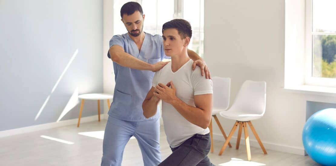 A patient is half-bending one leg while bringing his hands together at his core, demonstrating focus and balance. A physiotherapist stands beside him, providing guidance and support in a well-equipped physiotherapy clinic. The atmosphere is encouraging and professional, highlighting a collaborative approach to recovery.