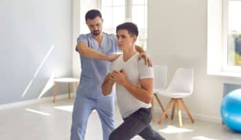 A patient is half-bending one leg while bringing his hands together at his core, demonstrating focus and balance. A physiotherapist stands beside him, providing guidance and support in a well-equipped physiotherapy clinic. The atmosphere is encouraging and professional, highlighting a collaborative approach to recovery.